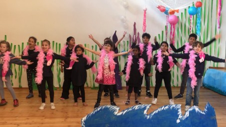 Eine Klasse mit 12 Kindern, schwarz gekleidet mit rosa Federboas und rosa Flamingoköpfen tanzt fröhlich mit erhobenen Händen einen Tanz. Die Meereskulisse ist gut zu sehen: Fische aus Luftballons in einem Netz, Algen an der Wand und im Vordergrund hohe Wellen aus Pappe.