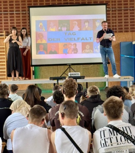 Im Vordergrund sitzen mehrere Schüler:innen. Die Gesichter der Schüler*innen sind nach vorne gerichtet, wo sich eine Leinwand befindet auf der steht: „Tag der Vielfalt“. Links auf dem Bild stehen zwei Lehrkräfte auf einer Bühne, eine davon dolmetscht in Gebärdensprache. Am rechten Rand der Bühne ist der Schullehrer Herr Eicker und hält ein Mikrofon in der Hand.