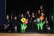 Auf einer Bühne sitzen Kinder und Lehrer des Workshops Schwarzlicht. Sie haben alle schwarze Kleidung an. Vor ihnen stehen zwei Blumen, die im Schwarzlicht leuchten.