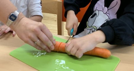 Ein Kind schneidet eine Möhre. Eine größere Hand hält die Möhre zusätzlich fest.