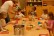 Fünf Kinder (von links nach rechts: Mädchen, Junge, Junge, Mädchen, Mädchen) sitzen um einen Tisch herum und belegen Hotdogs. Die Zutaten stehen vor ihnen auf dem Tisch bereit. Ein hoher Edelstahltopf mit Würstchen, Weidenkörbchen mit Brötchen, Glasschälchen mit sauren Gurken sowie ein Glasschälchen mit Röstzwiebeln.  Außerdem stehen eine Flasche Senf und eine Flasche Ketchup bereit. Als Getränke stehen zwei Flaschen Wasser und eine Kanne Tee auf dem Tisch. Eine Frau hilft einem Jungen in der Mitte beim Auftragen des  Ketchups.

