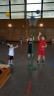 Drei Jungen stehen um einen Basketballkorb herum und versuchen ihn mit Softbällen zu treffen. Ein Mädchen kommt von etwas weiter weg auf den Basketballkorb zugelaufen.