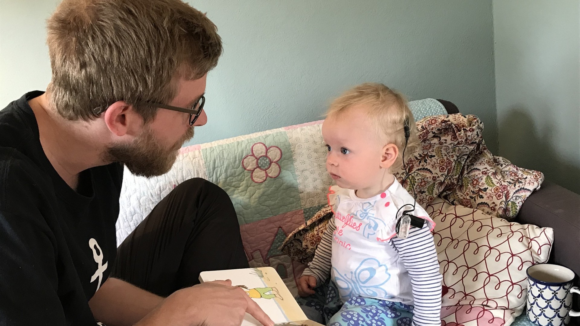 Ein Mann und ein Kleinkind, das ein Cochlea-Implantat trägt, sitzen gemeinsam auf einem Sofa und betrachten konzentriert ein Bilderbuch im Rahmen der häuslichen Frühförderung.