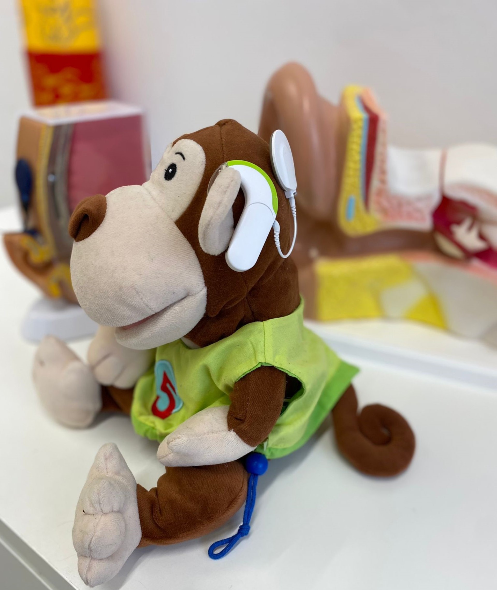 Die Plüschfigur eines Affen, das Maskottchen „Gerry“, trägt ein weißes Cochlea-Implantat (CI) am Kopf, um Kindern spielerisch den Umgang mit Hörtechnik zu vermitteln; im Hintergrund sind anatomische Modelle des menschlichen Ohrs zu sehen.
