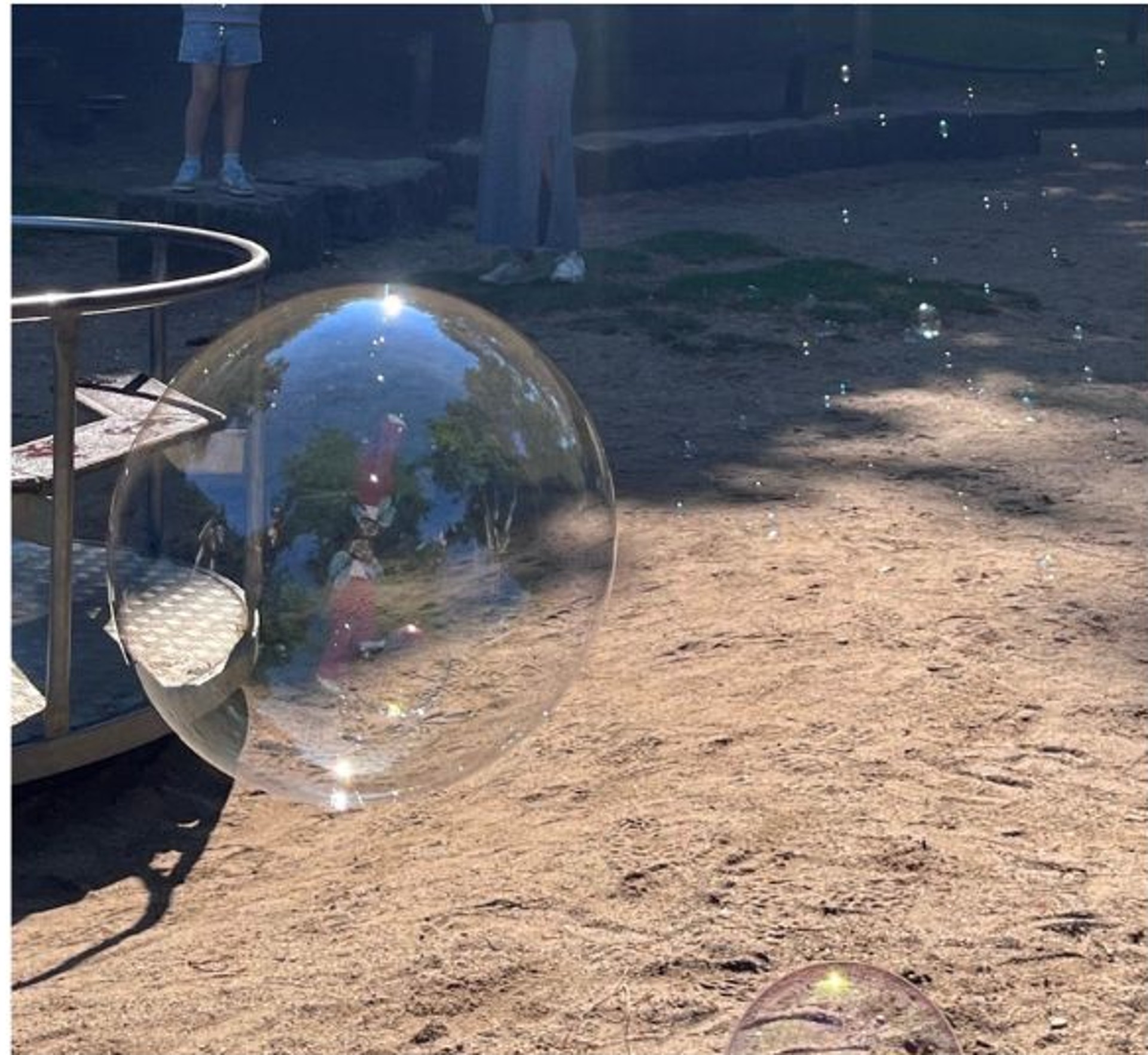 Ein Spielgerät auf dem Spielplatz zeigt auf seiner Außenseite einen rot- weißen Pilz. Dahinter können Kleinkinder über ein paar Stufen auf ein Podest klettern. Rechts neben dem Pilz führt die kurze Rutsche ab. Im Hintergrund ist die längere Rutsche eines anderen Klettergerüsts zu sehen. Beide Spielgeräte stehen auf Sandboden. Im Hintergrund sind grüne Bäume zu sehen.