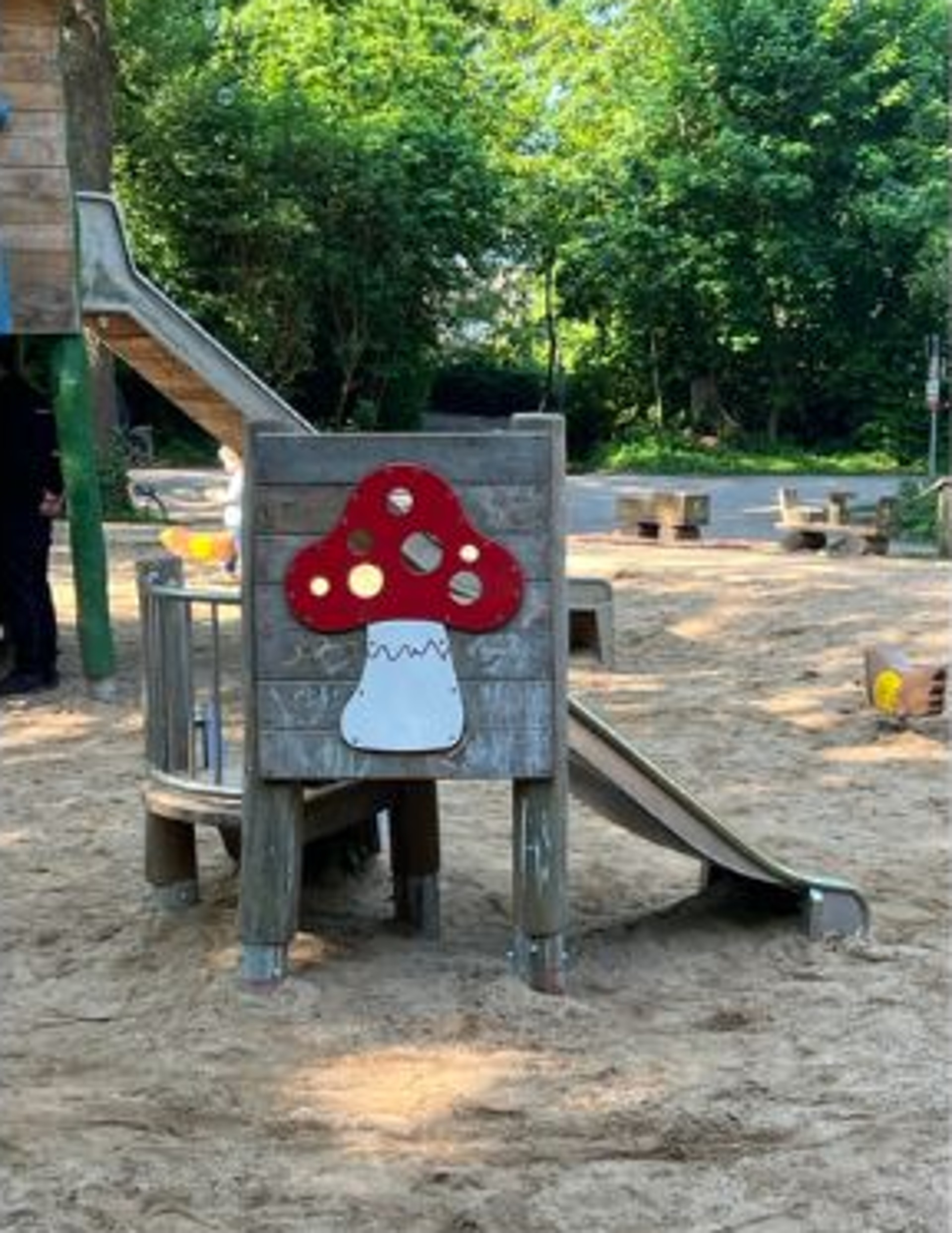 Ein Spielgerät auf dem Spielplatz zeigt auf seiner Außenseite einen rot- weißen Pilz. Dahinter können Kleinkinder über ein paar Stufen auf ein Podest klettern. Rechts neben dem Pilz führt die kurze Rutsche ab. Im Hintergrund ist die längere Rutsche eines anderen Klettergerüsts zu sehen. Beide Spielgeräte stehen auf Sandboden. Im Hintergrund sind grüne Bäume zu sehen.