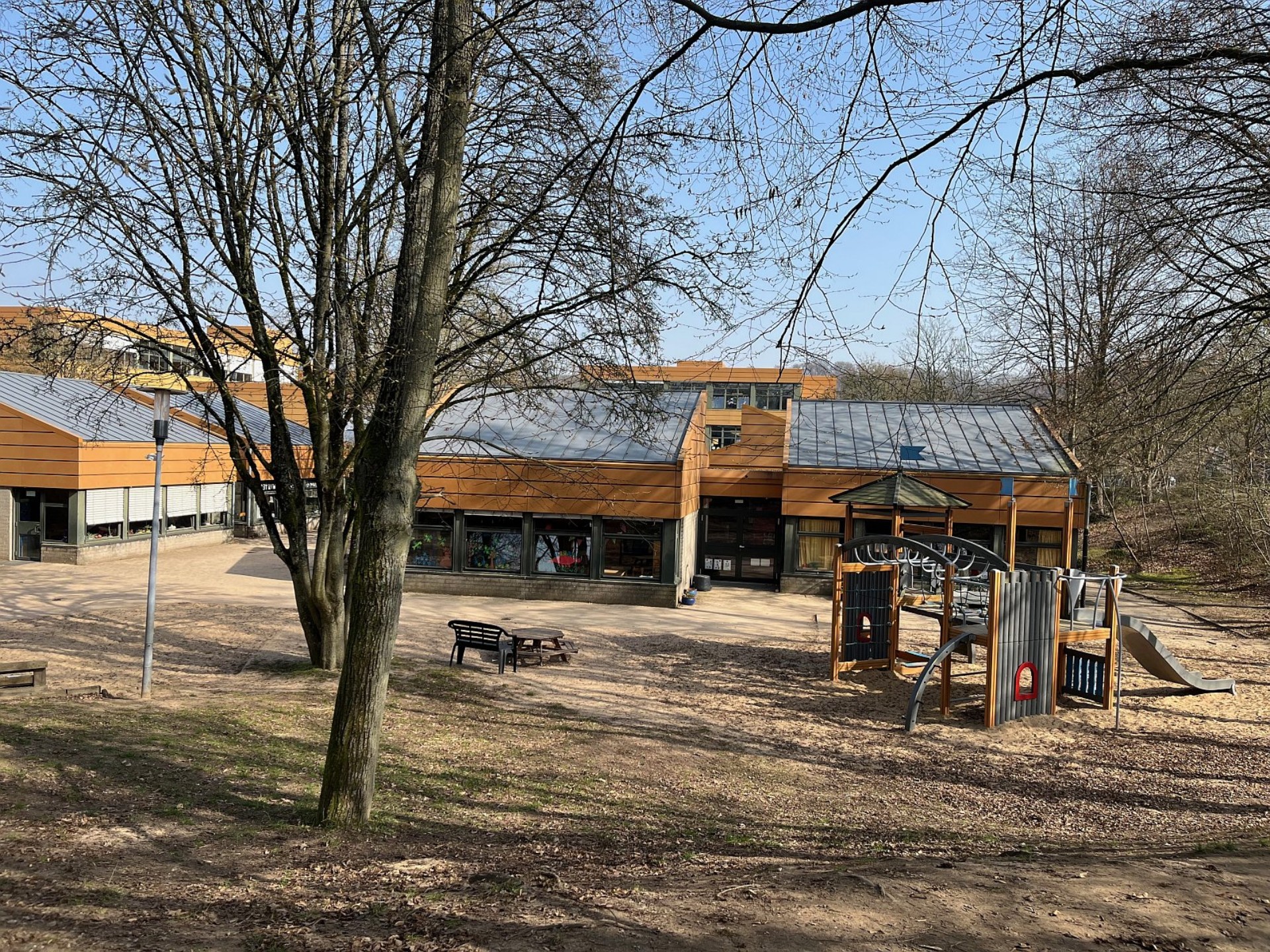 Außenbereich des Kindergartens mit einem Spielgerüst, einer Sitzgruppe und flachen Schulgebäuden, eingebettet in eine bewaldete Umgebung.