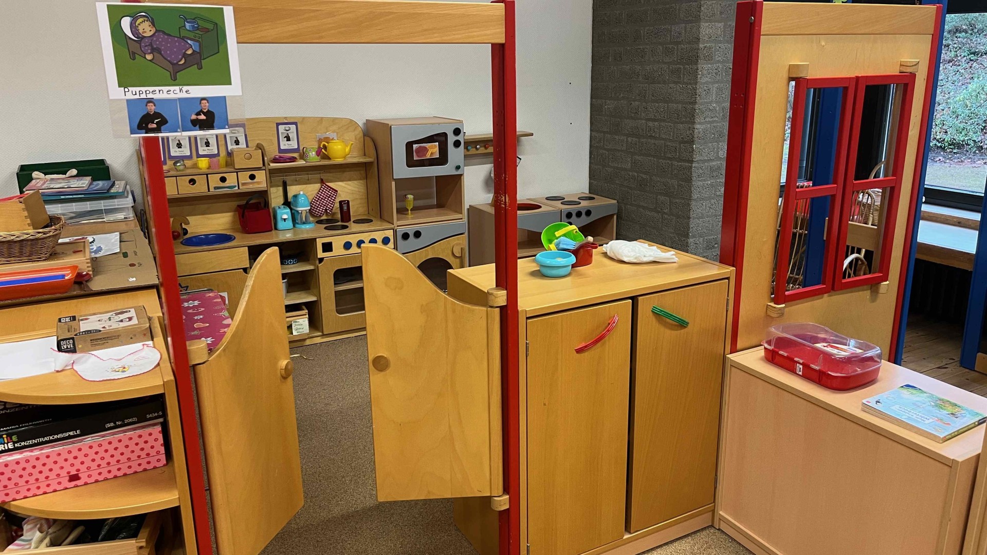 Spielecke für Kinder aus hellem Holz. Im Zentrum steht eine kleine Spielküche mit Herd, Mikrowelle und Zubehör. Der Bereich ist durch niedrige Holzelemente und eine kleine Schwingtür begrenzt. Ein Schild über dem Eingang zeigt das Wort „Puppenecke“ kombiniert mit Gebärden-Symbolen.