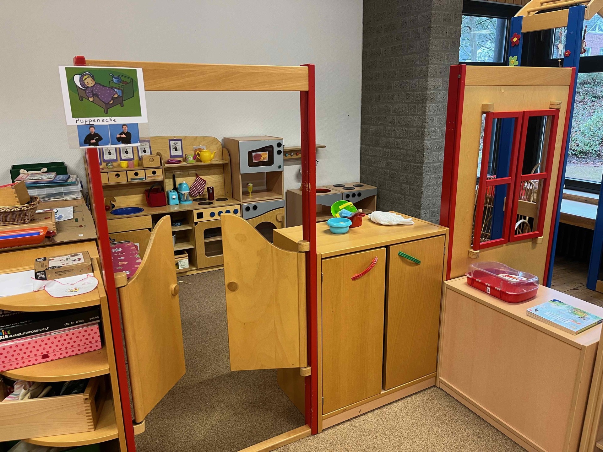 Spielecke für Kinder aus hellem Holz. Im Zentrum steht eine kleine Spielküche mit Herd, Mikrowelle und Zubehör. Der Bereich ist durch niedrige Holzelemente und eine kleine Schwingtür begrenzt. Ein Schild über dem Eingang zeigt das Wort „Puppenecke“ kombiniert mit Gebärden-Symbolen.
