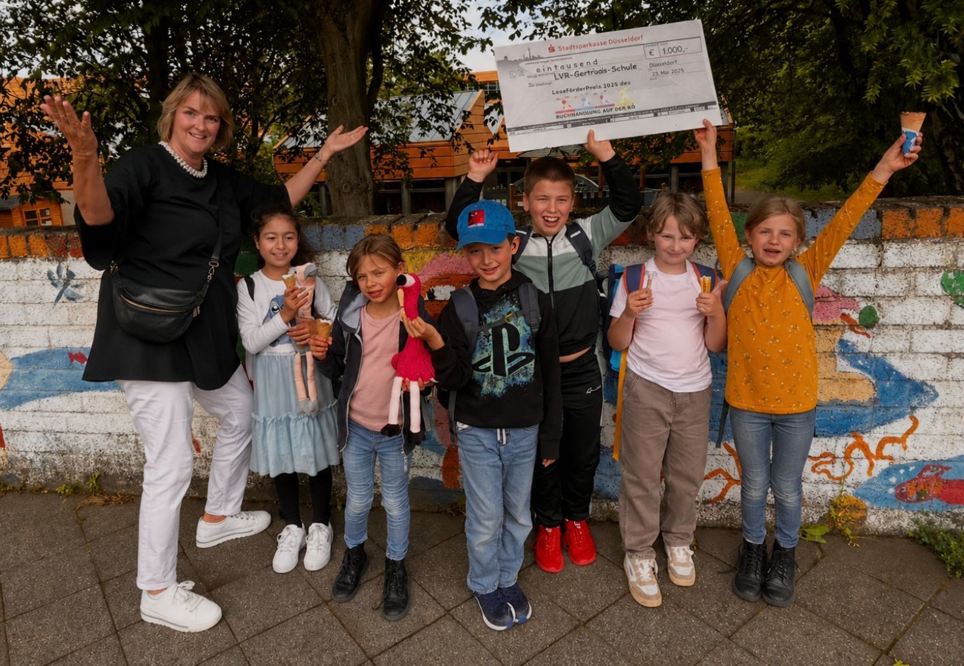 Ein Spielgerät auf dem Spielplatz zeigt auf seiner Außenseite einen rot- weißen Pilz. Dahinter können Kleinkinder über ein paar Stufen auf ein Podest klettern. Rechts neben dem Pilz führt die kurze Rutsche ab. Im Hintergrund ist die längere Rutsche eines anderen Klettergerüsts zu sehen. Beide Spielgeräte stehen auf Sandboden. Im Hintergrund sind grüne Bäume zu sehen.