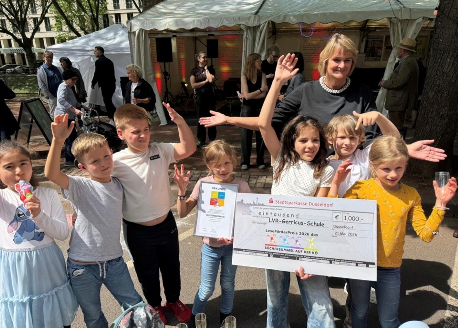 Eine Gruppe von acht Personen steht auf dem Festgelände und hält einen symbolischen Scheck der Stadtsparkasse Düsseldorf über 1000 € in die Kamera. Der Scheck ist an die „LVR-Gerricus-Schule“ adressiert. Im Hintergrund sind verschiedene Festzelte und andere Personen zu sehen.