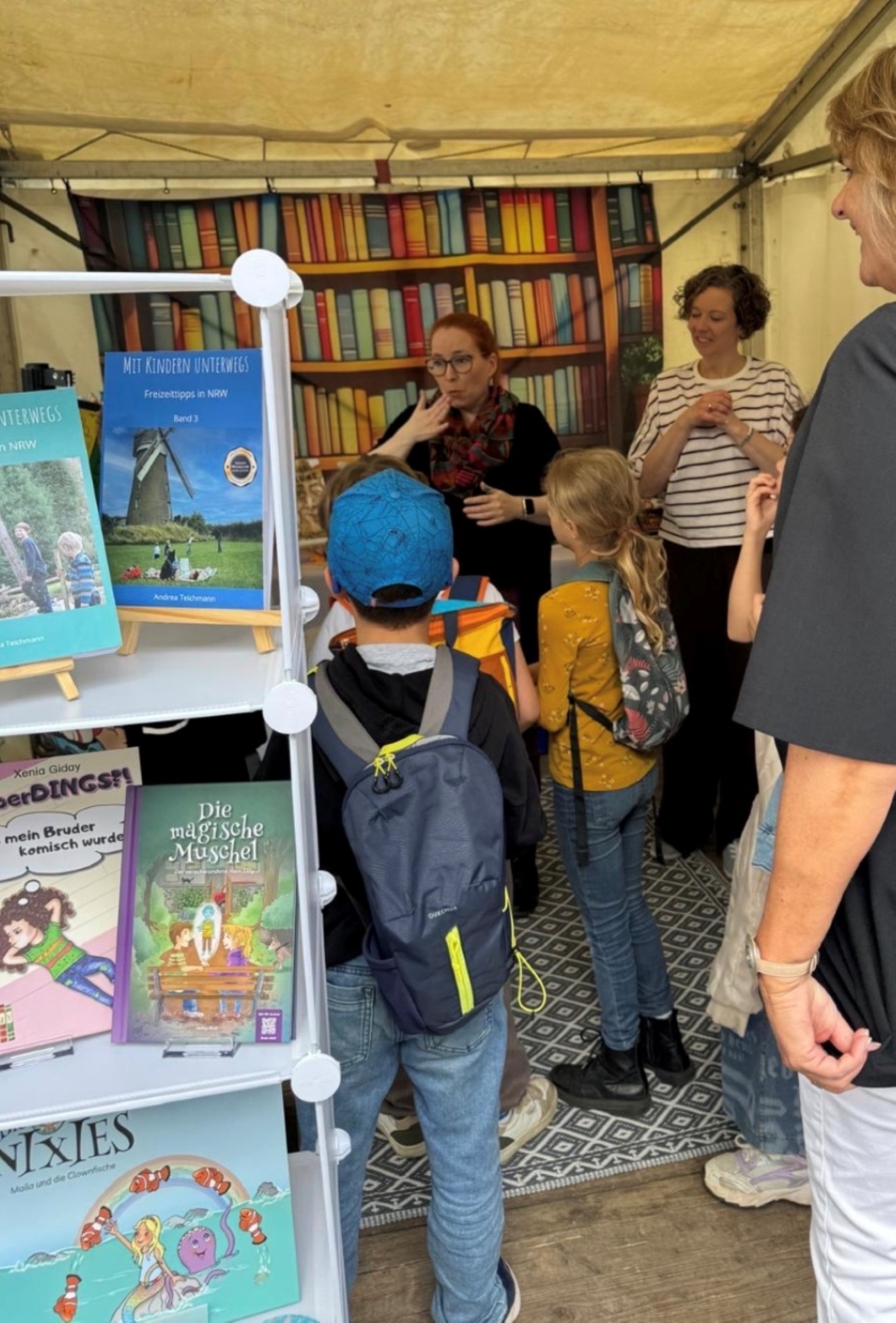Es sind fünf Personen an einem Bücherstand mit Kinderbüchern zu sehen. Eine Frau erzählt etwas und eine Frau gebärdet. Die anderen Personen hören zu. Im Vordergrund steht ein Regal mit verschiedenen Kinderbüchern.