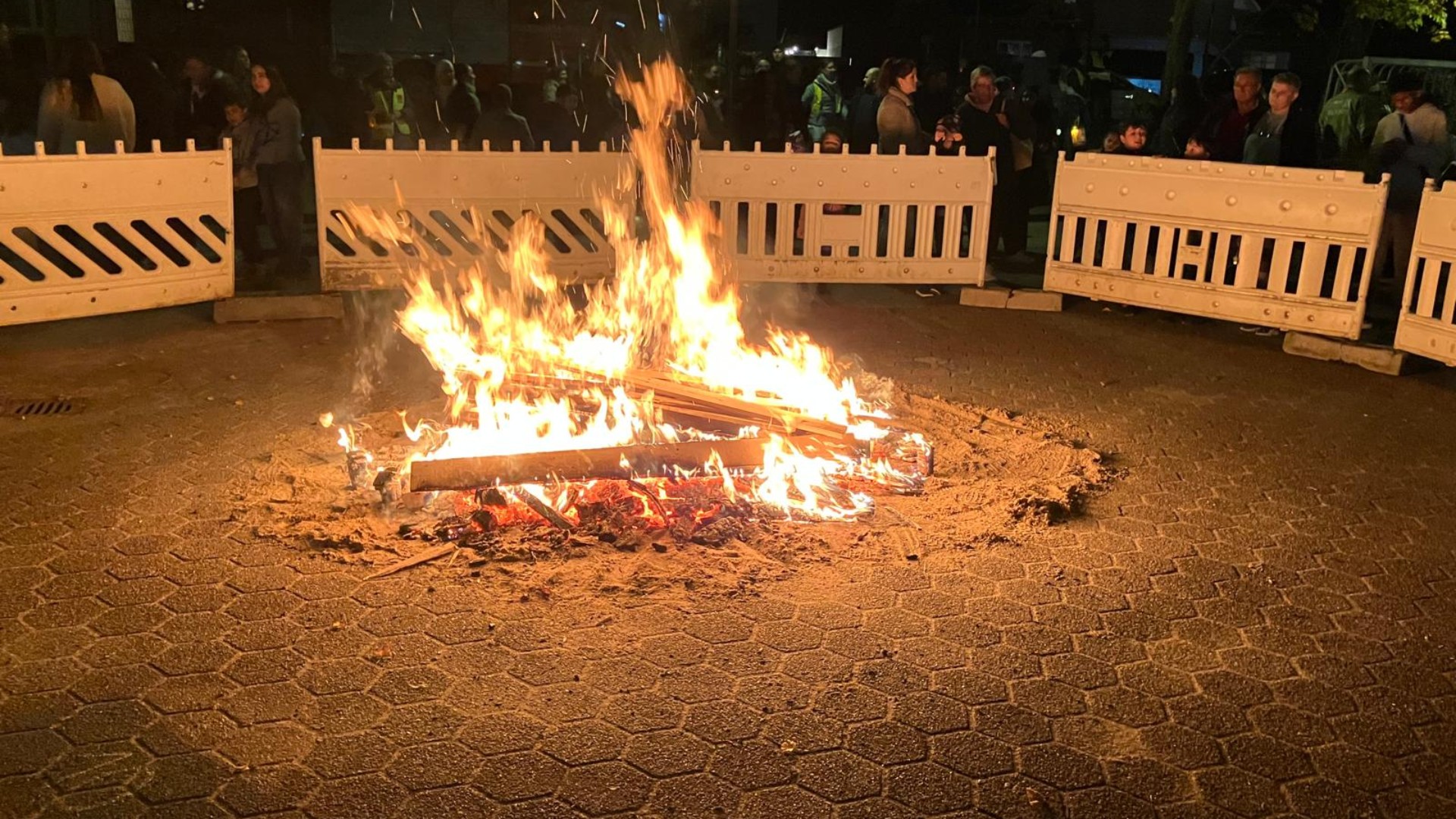 Ein loderndes Martinsfeuer brennt am Abend auf einem gepflasterten Platz. Das Feuer ist kreisförmig mit Sand unterlegt und durch weiße Absperrgitter gesichert. Im Hintergrund stehen im Halbdunkeln zahlreiche Menschen – Kinder und Erwachsene –, die das Spektakel beobachten. Funken sprühen in den Nachthimmel.