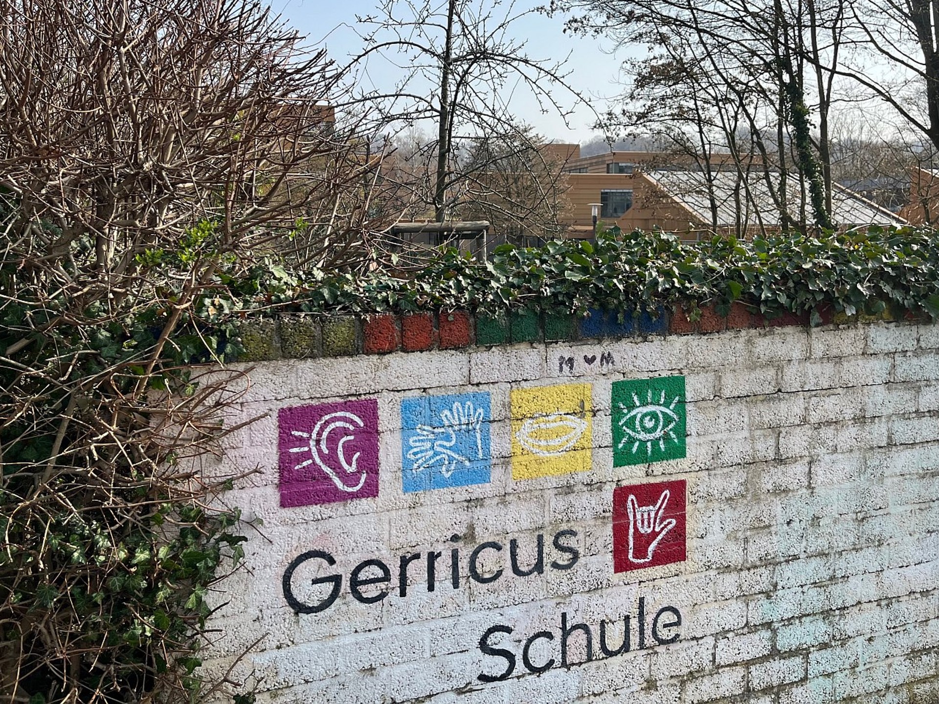 Weiße Mauer mit dem bunten Logo der Gerricus-Schule und Symbolen für Hören, Gebärden und Sehen. Dahinter sind Bäume und die Fassade der LVR-Schule in Düsseldorf zu sehen.