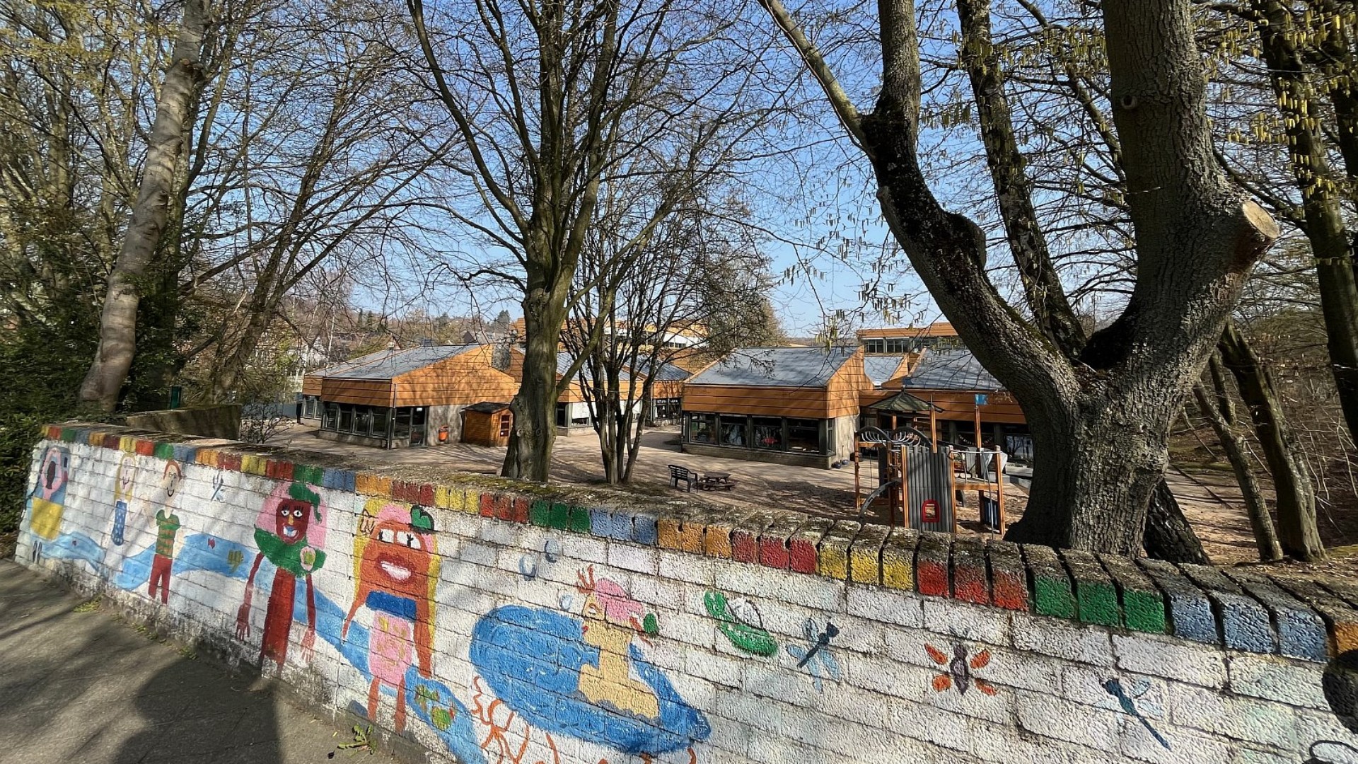 Eine Nahaufnahme eines Materialregals vor einer lila-gelben Wand. Auf orangefarbenen Tabletts stehen zahlreiche Gläser mit Pinseln, Farbmischpaletten und gebrauchte Schwämme.