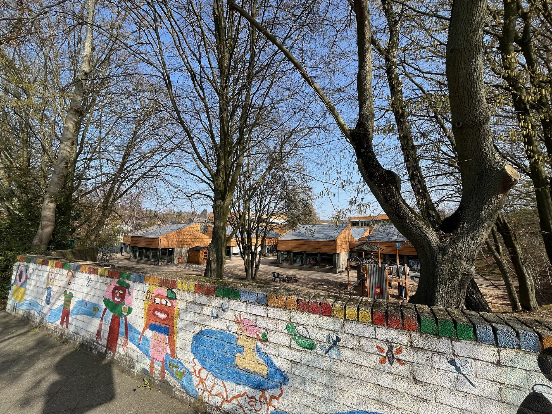 Blick über eine bunt bemalte Mauer auf das Schulgelände mit flachen Gebäuden, hohen, noch kahlen Bäumen und einem Spielplatzbereich im Hintergrund.