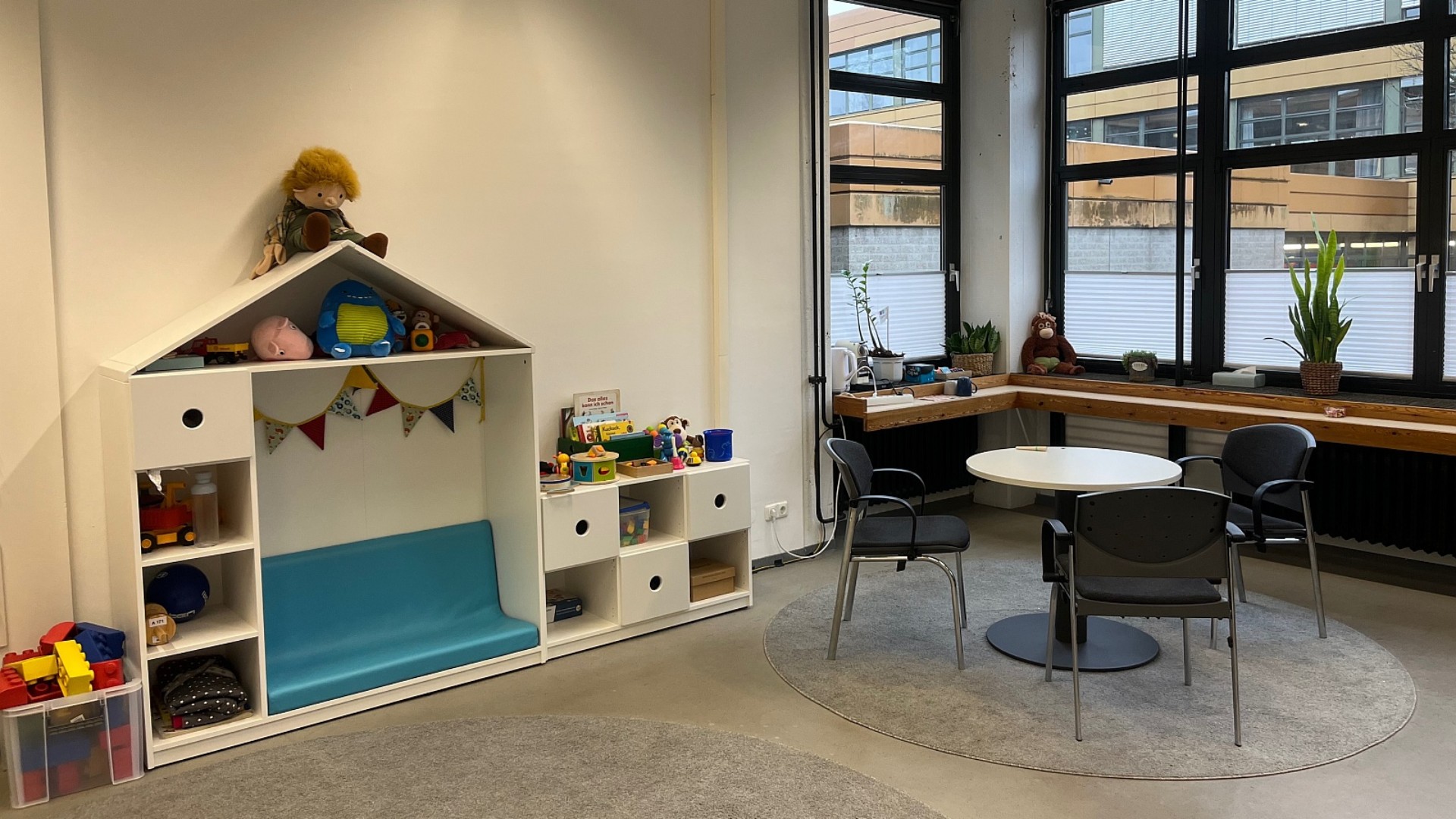 Ein Raum  mit einem weißen Regal mit Spielsachen wie Plüschtieren und Büchern und einem runden Tisch mit drei Stühlen.