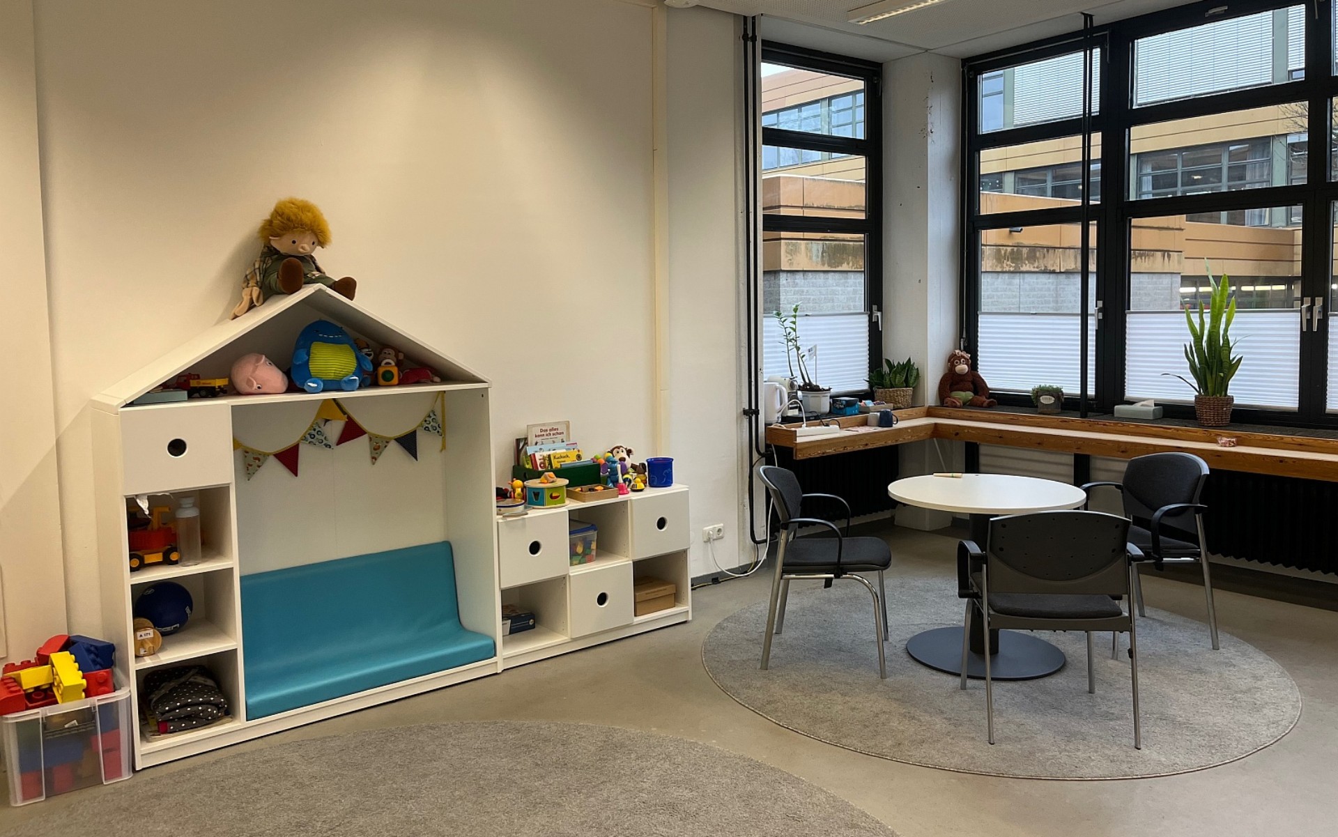 Ein Raum  mit einem weißen Regal mit Spielsachen wie Plüschtieren und Büchern und einem runden Tisch mit drei Stühlen.