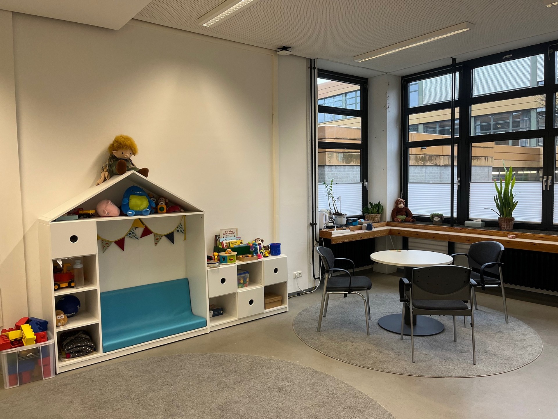 Ein Raum  mit einem weißen Regal mit Spielsachen wie Plüschtieren und Büchern und einem runden Tisch mit drei Stühlen.