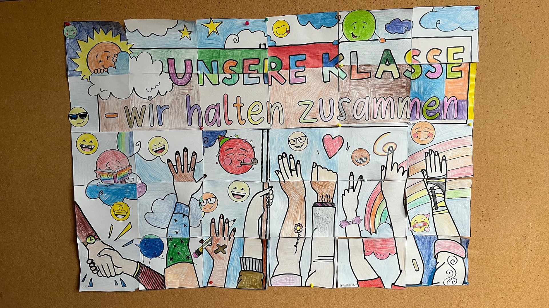 Ein großes, buntes selbstgezeichnetes Bild hängt an einer Korkwand: Darauf ist der Schriftzug zu lesen "Unsere Klasse – wir halten zusammen" und mehrere Arme und Hände, die wie bei einer Demonstration in die Höhe gehalten werden.