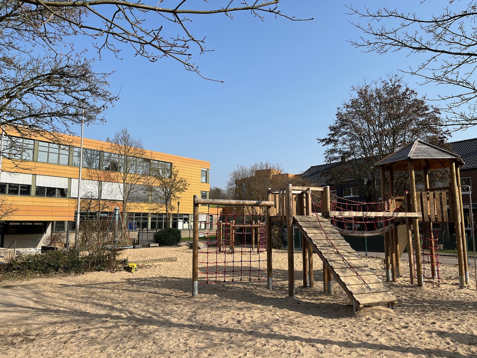 Spielplatz der Grundschule mit einem großen Holz-Klettergerüst im Sandkasten, dahinter das moderne, gelb-orangefarbene Schulgebäude der LVR-Gerricus-Schule.