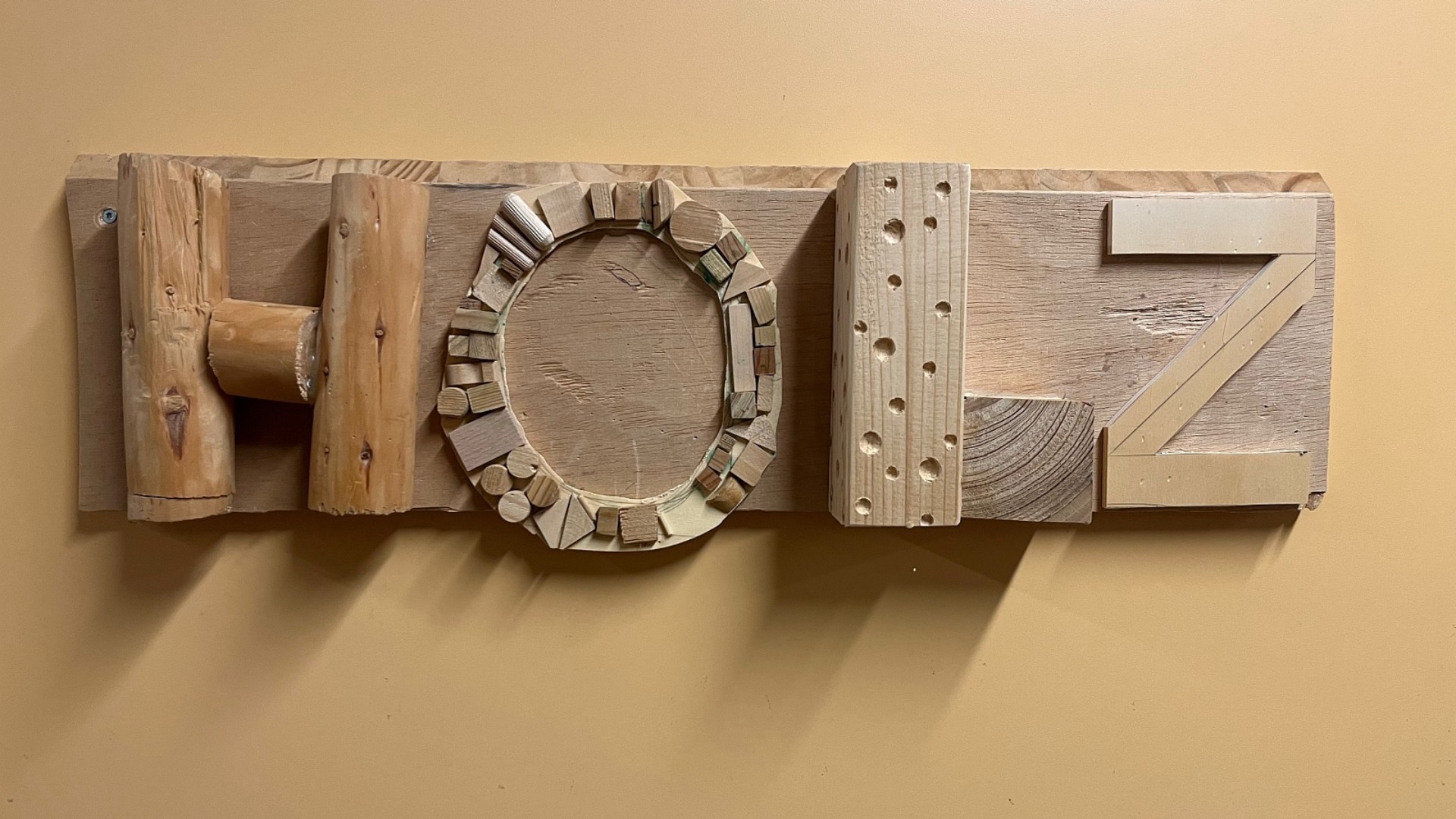 Ein hölzernes Schild an einer schlichten, ockerfarbenen Wand zeigt das Wort „HOLZ“. Das Besondere an diesem Schild ist, dass jeder Buchstabe aus verschiedenen Holzresten und mit unterschiedlichen Techniken gestaltet wurde.