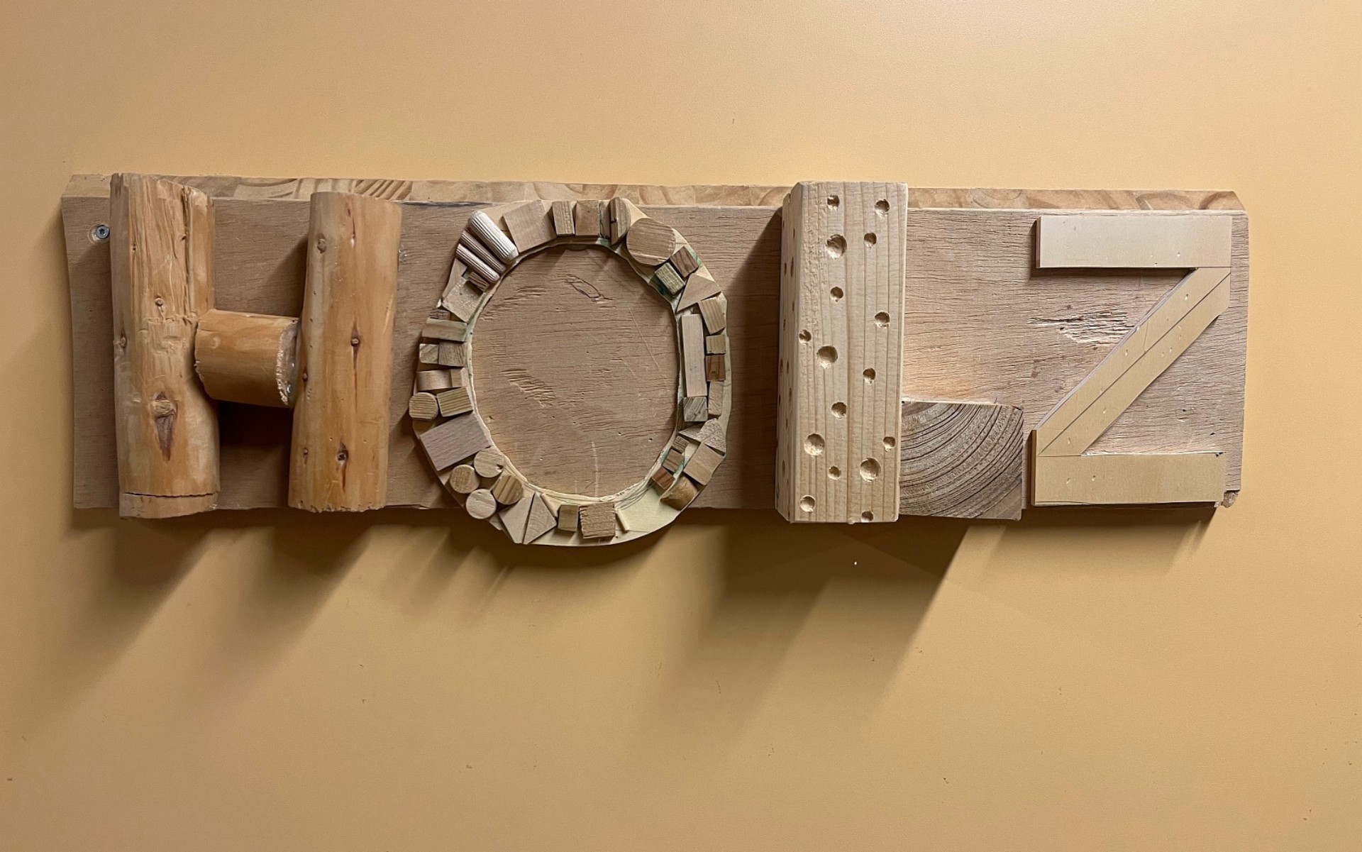 Ein hölzernes Schild an einer schlichten, ockerfarbenen Wand zeigt das Wort „HOLZ“. Das Besondere an diesem Schild ist, dass jeder Buchstabe aus verschiedenen Holzresten und mit unterschiedlichen Techniken gestaltet wurde.