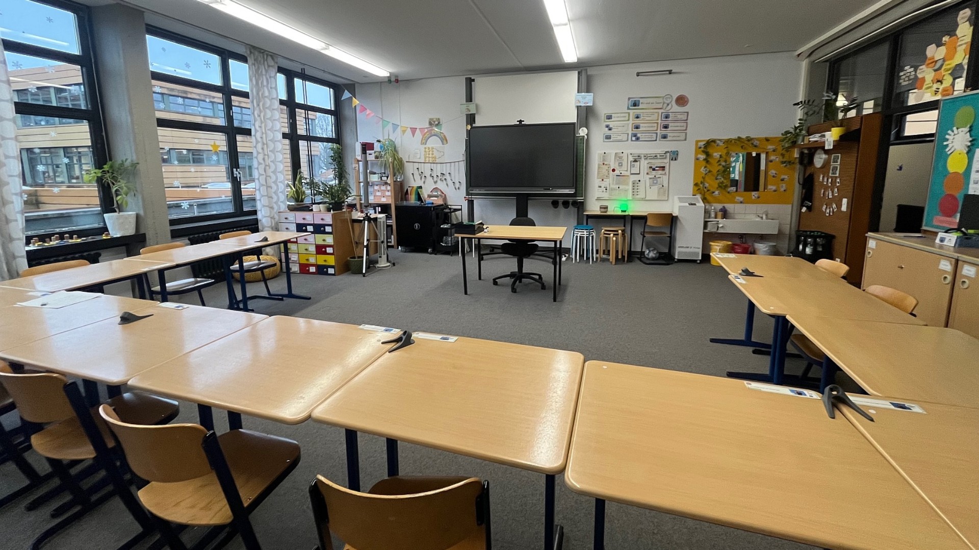 Ein helles Klassenzimmer mit grauen Teppichboden, das in einer Hufeisenform (U-Form) mit hellen Holztischen arrangiert ist.  Sitzordnung: Die Einzeltische sind so zusammengestellt, dass sie eine große, offene Mitte bilden, die zum Lehrertisch und einem großen digitalen Whiteboard an der Stirnseite ausgerichtet ist.  Ausstattung: Im hinteren Bereich befinden sich verschiedene Schränke, ein Waschbecken mit gelben Akzenten, eine bunte Kommode mit vielen Schubladen und diverse Lernmaterialien an den Wänden.