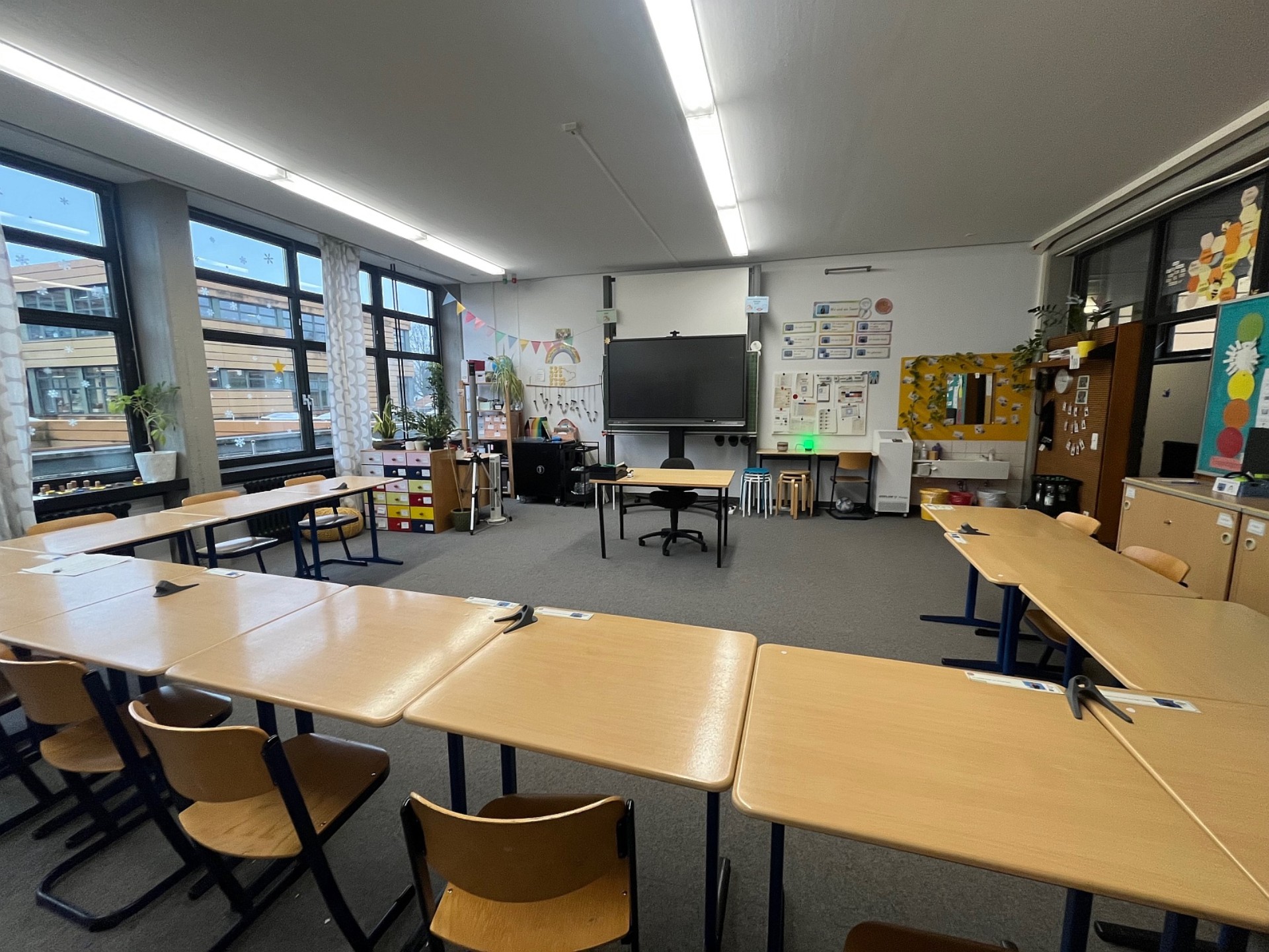 Ein helles Klassenzimmer mit grauen Teppichboden, das in einer Hufeisenform (U-Form) mit hellen Holztischen arrangiert ist.  Sitzordnung: Die Einzeltische sind so zusammengestellt, dass sie eine große, offene Mitte bilden, die zum Lehrertisch und einem großen digitalen Whiteboard an der Stirnseite ausgerichtet ist.  Ausstattung: Im hinteren Bereich befinden sich verschiedene Schränke, ein Waschbecken mit gelben Akzenten, eine bunte Kommode mit vielen Schubladen und diverse Lernmaterialien an den Wänden.