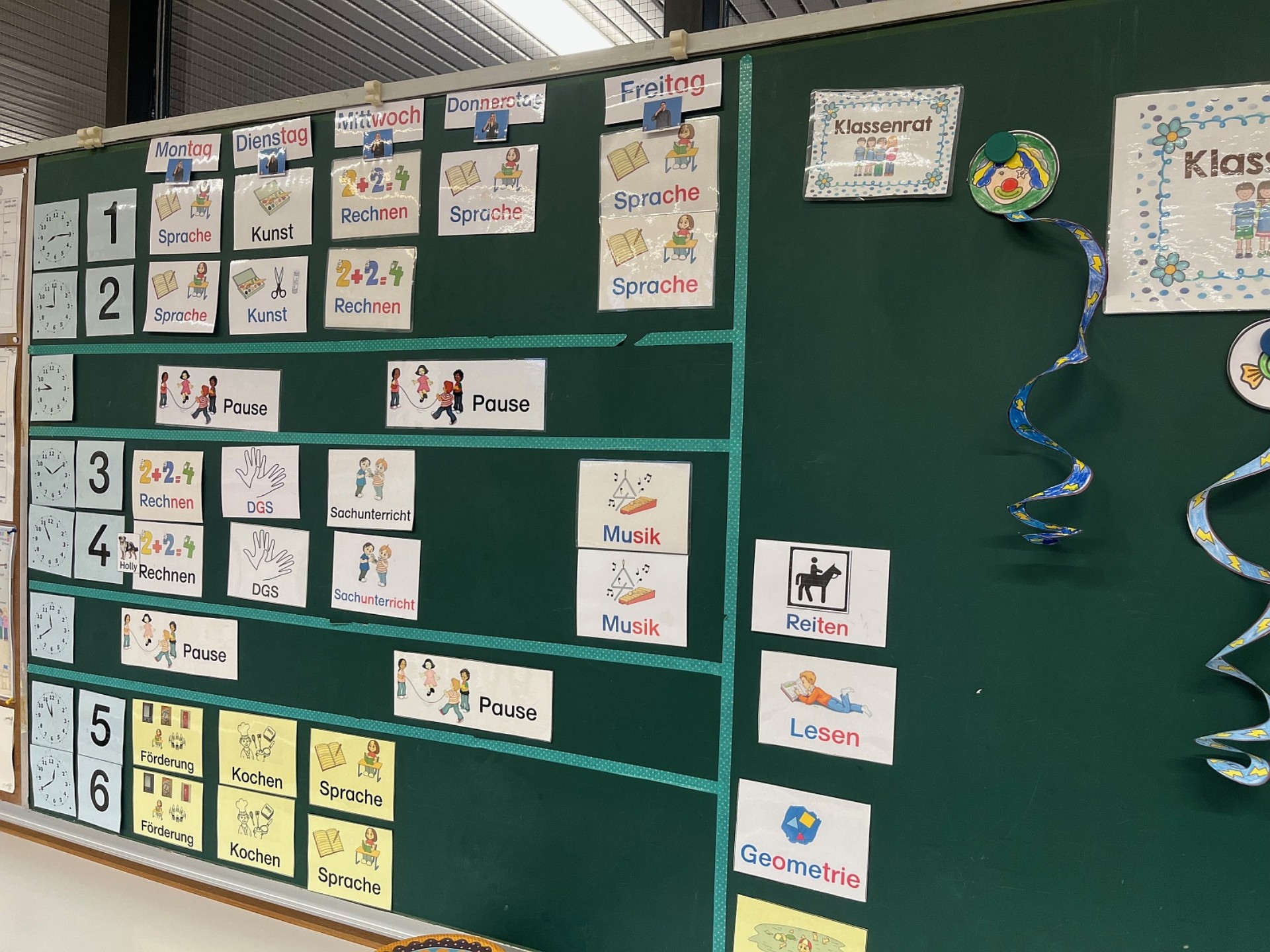 Eine grüne Kreidetafel, die als visueller Stundenplan genutzt wird und mit magnetischen Karten strukturiert ist.  Zeitliche Gliederung: Am oberen Rand sind die Wochentage von Montag bis Freitag beschriftet. Am linken Rand markieren Ziffern (1 bis 6) und kleine Uhrensymbole die Unterrichtsstunden.  Visualisierung: Die einzelnen Fächer wie „Sprache“, „Rechnen“, „Kunst“ oder „Sachunterricht“ sind durch farbige Karten mit Piktogrammen dargestellt.  Besonderheiten: Es gibt spezielle Karten für „DGS“ (Deutsche Gebärdensprache), „Förderung“, „Kochen“ und „Reiten“. Die Pausenzeiten sind durch lange weiße Streifen mit spielenden Kindern deutlich hervorgehoben.  Zusatzbereiche: Auf der rechten Tafelseite befinden sich Überschriften wie „Klassenrat“ sowie Symbole für freie Lernzeiten oder individuelle Aufgaben wie „Lesen“ und „Geometrie“.