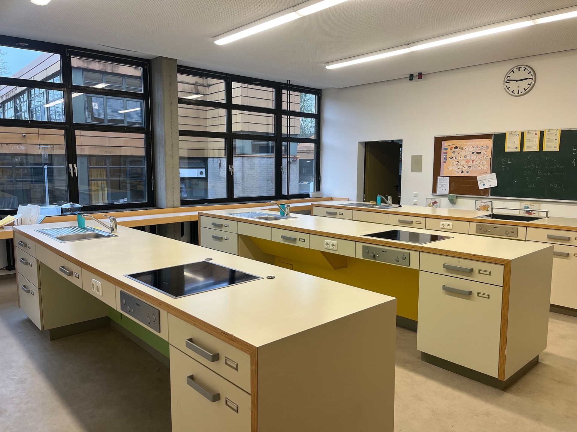 Eine moderne, helle Lehrküche mit mehreren freistehenden Kochinseln. Jede Station verfügt über ein Induktionskochfeld, ein Spülbecken und integrierte Unterschränke in Beige mit gelben oder grünen Akzenten. Im Hintergrund befinden sich eine große Fensterfront sowie eine Tafel mit einem Rezept und einer analogen Wanduhr.