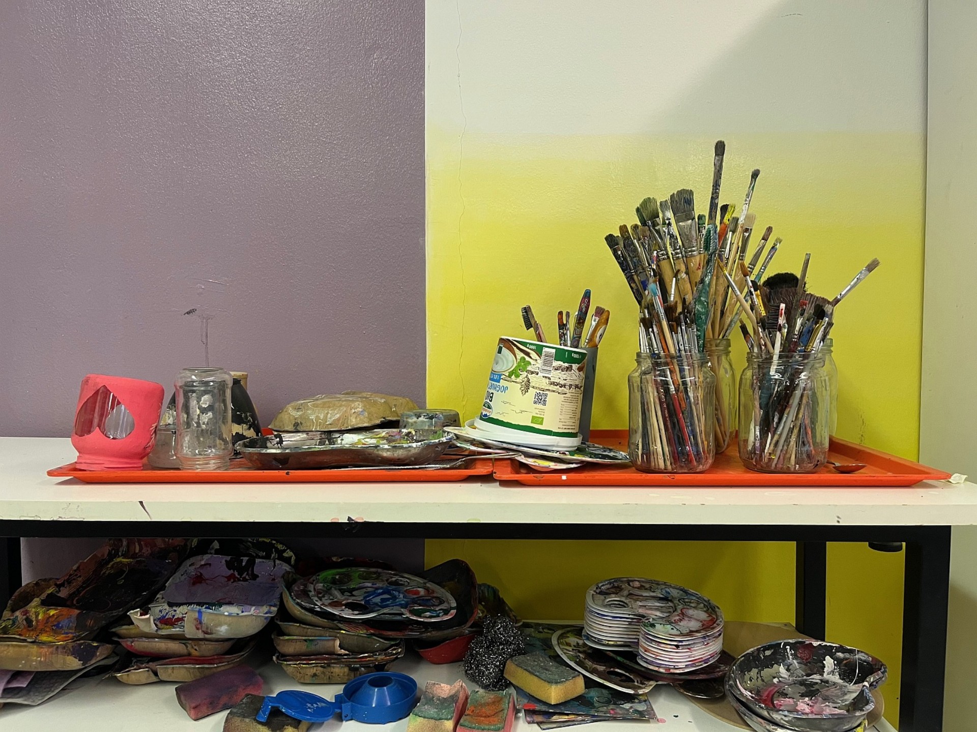 Eine Nahaufnahme eines Materialregals vor einer lila-gelben Wand. Auf orangefarbenen Tabletts stehen zahlreiche Gläser mit Pinseln, Farbmischpaletten und gebrauchte Schwämme.