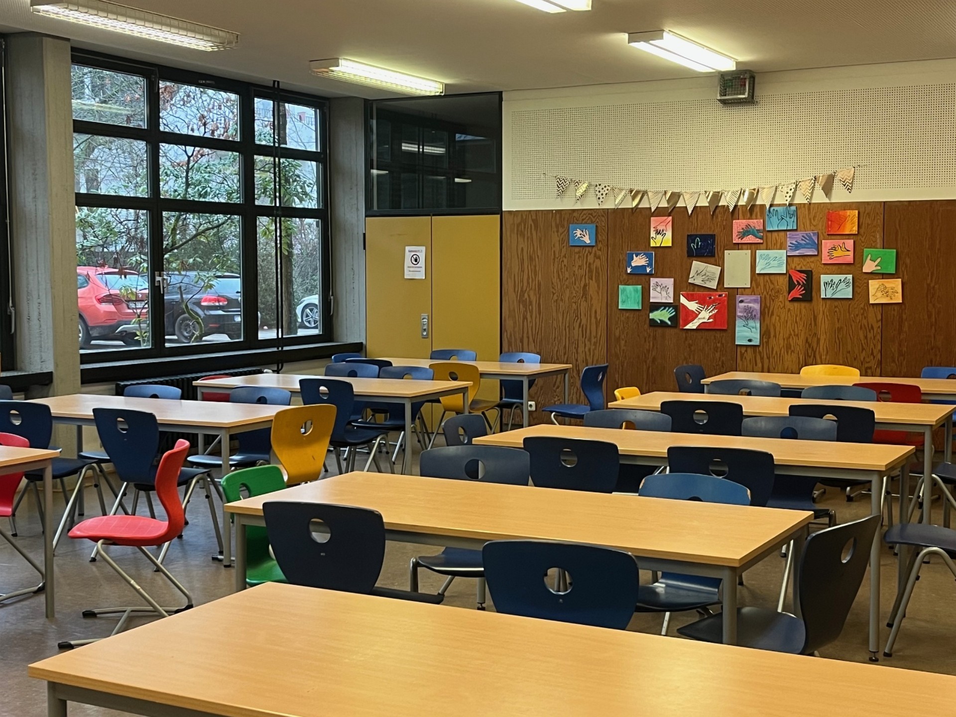Ein Speiseraum mit vielen Holztischen und bunten Stühlen. Die Holzwand im Hintergrund ist mit farbenfrohen Schülerarbeiten (Handabdrücken) und einer Wimpelkette dekoriert.