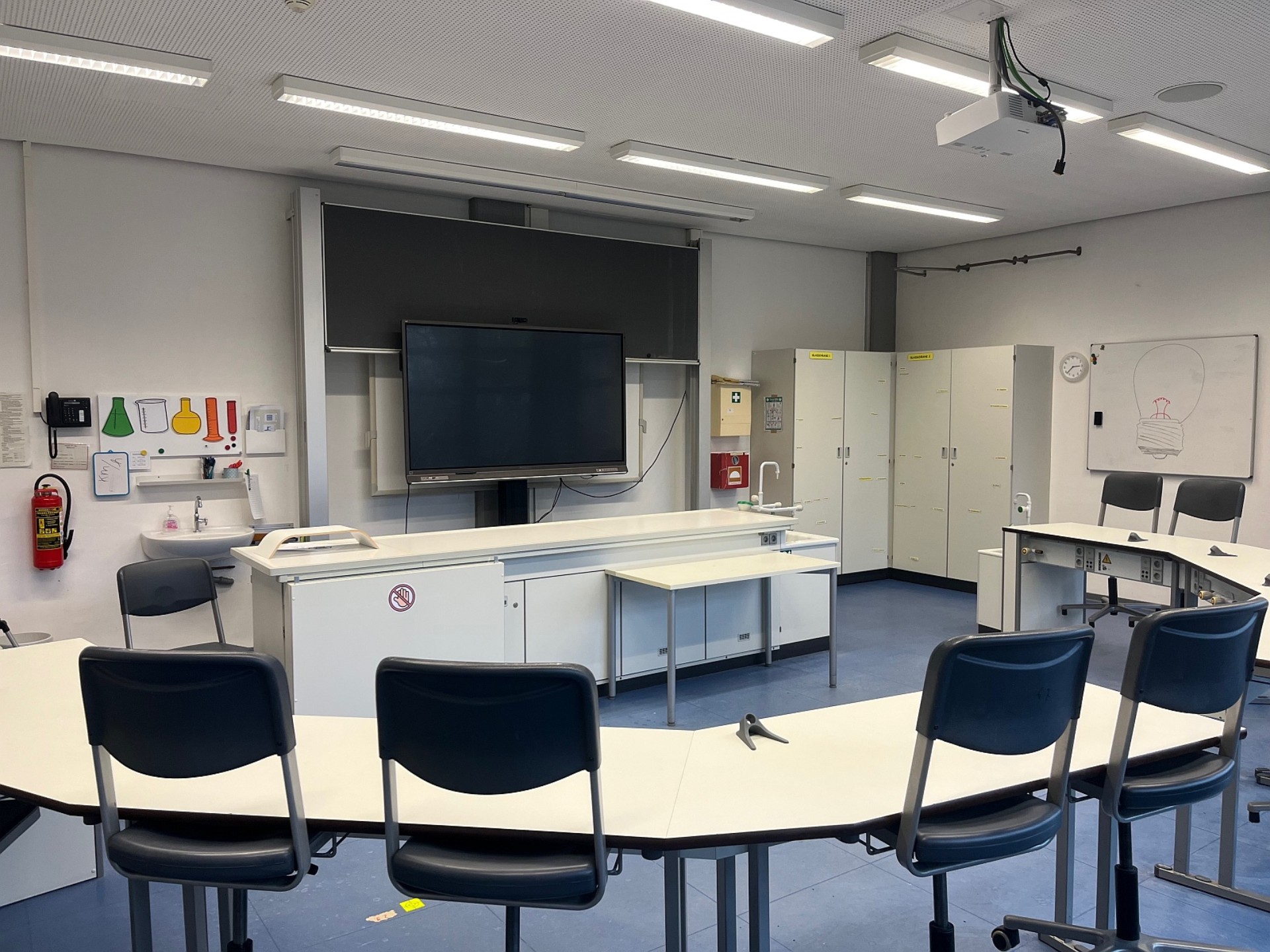 Ein moderner Fachraum mit weißen, halbkreisförmig angeordneten Labortischen. Im Zentrum stehen ein Experimentiertisch und ein großes digitales Whiteboard. Ein Waschbecken mit Gefahrensymbol-Piktogrammen und Sicherheitsschränke ergänzen die Ausstattung.