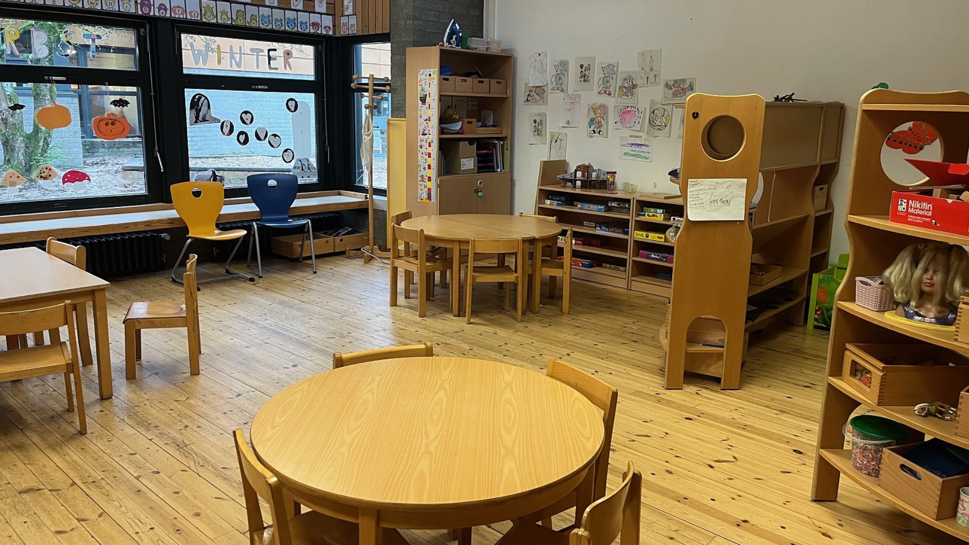 Ein freundlicher Raum für den Ganztag mit warmem Holzfußboden und runden Spieltischen. Niedrige Regale mit Brettspielen, eine Puppenecke und Fensterdekorationen schaffen eine wohnliche Atmosphäre.