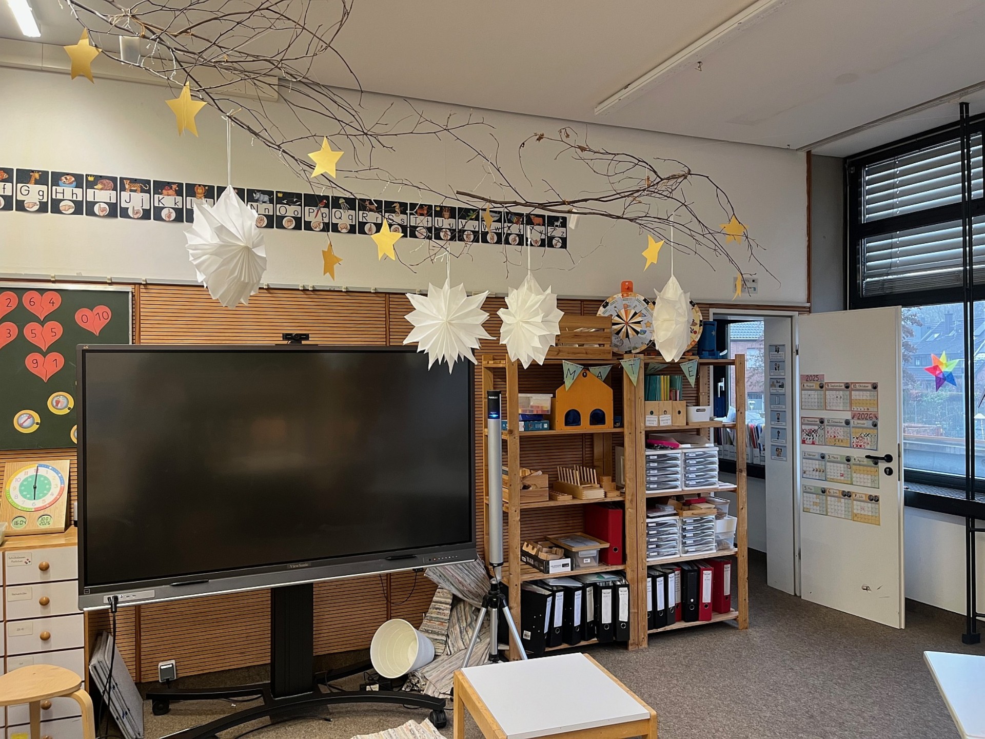 Ein gemütlicher Lernbereich mit einem digitalen Whiteboard und Regalsystemen. Ein großer Naturast, der mit Papiersternen und -lampen dekoriert ist, hängt von der Decke.