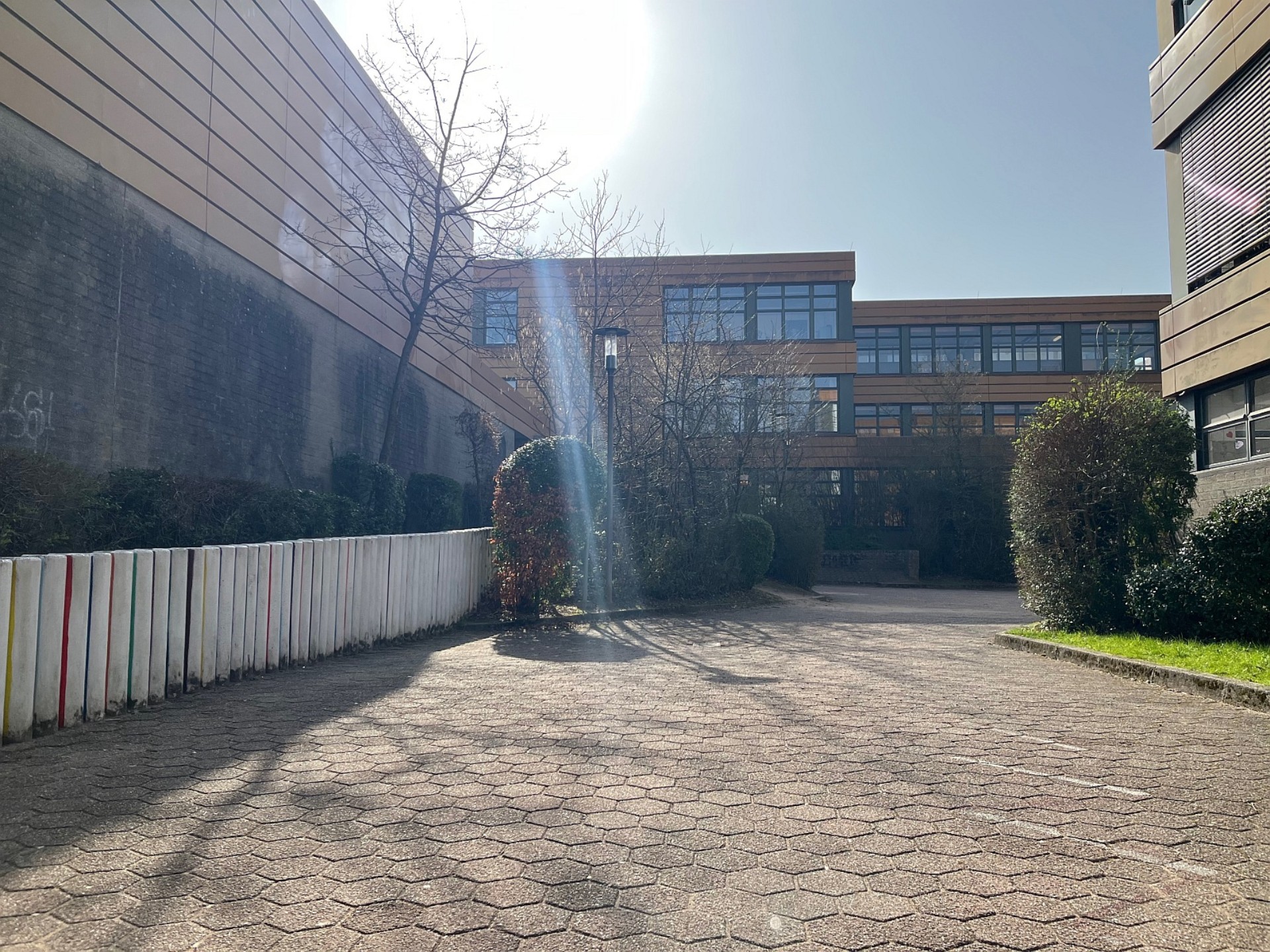 Gepflasterter Weg zwischen den Schulgebäuden der Sekundarstufe bei strahlendem Sonnenschein, flankiert von einer niedrigen, bunten Palisadenwand und Gebüschen.