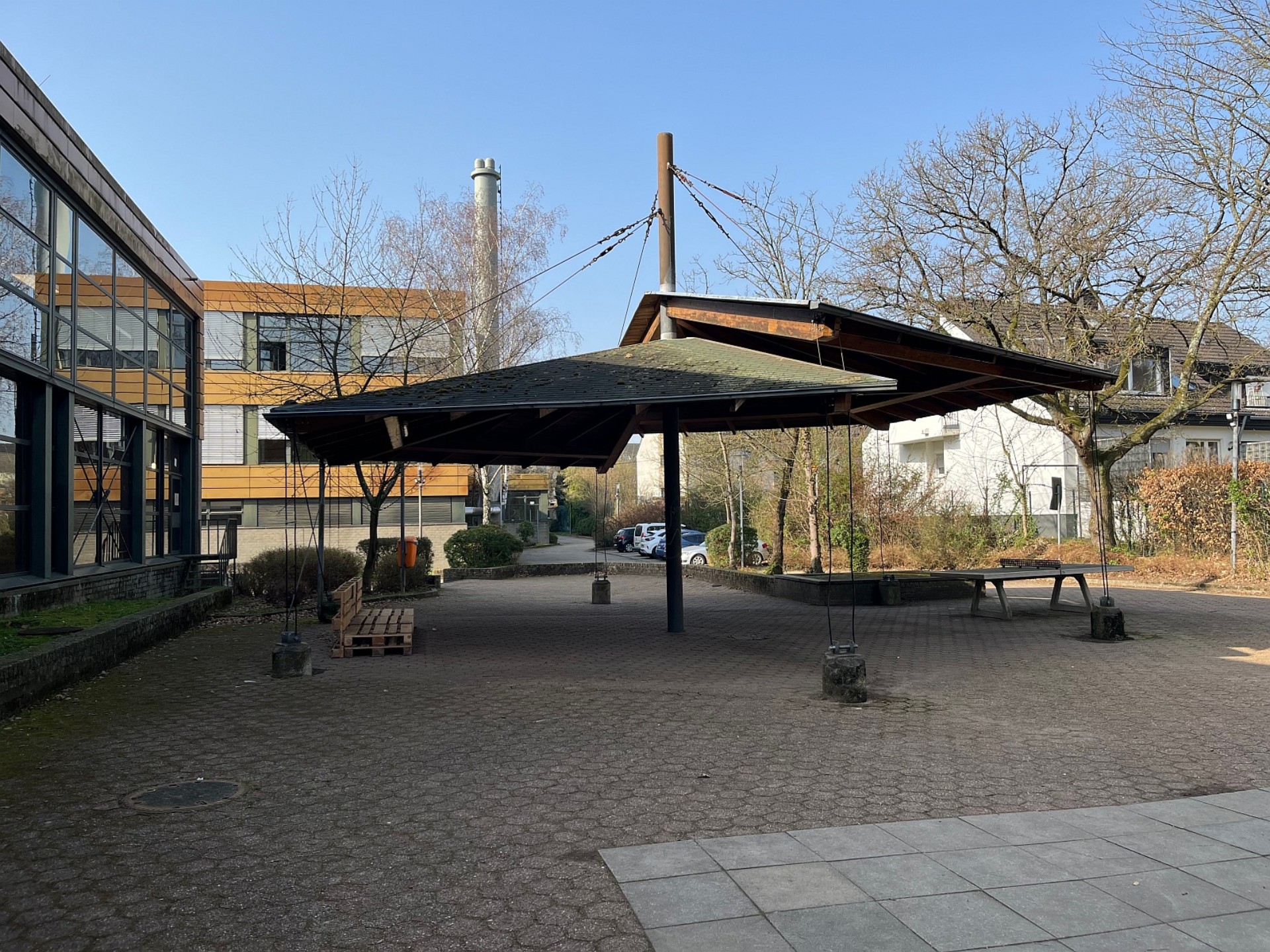 Gepflasterter Schulhof der Sekundarstufe mit einer großen, zeltartigen Holzüberdachung, einer Tischtennisplatte im Hintergrund und modernen Schulgebäuden unter blauem Himmel.