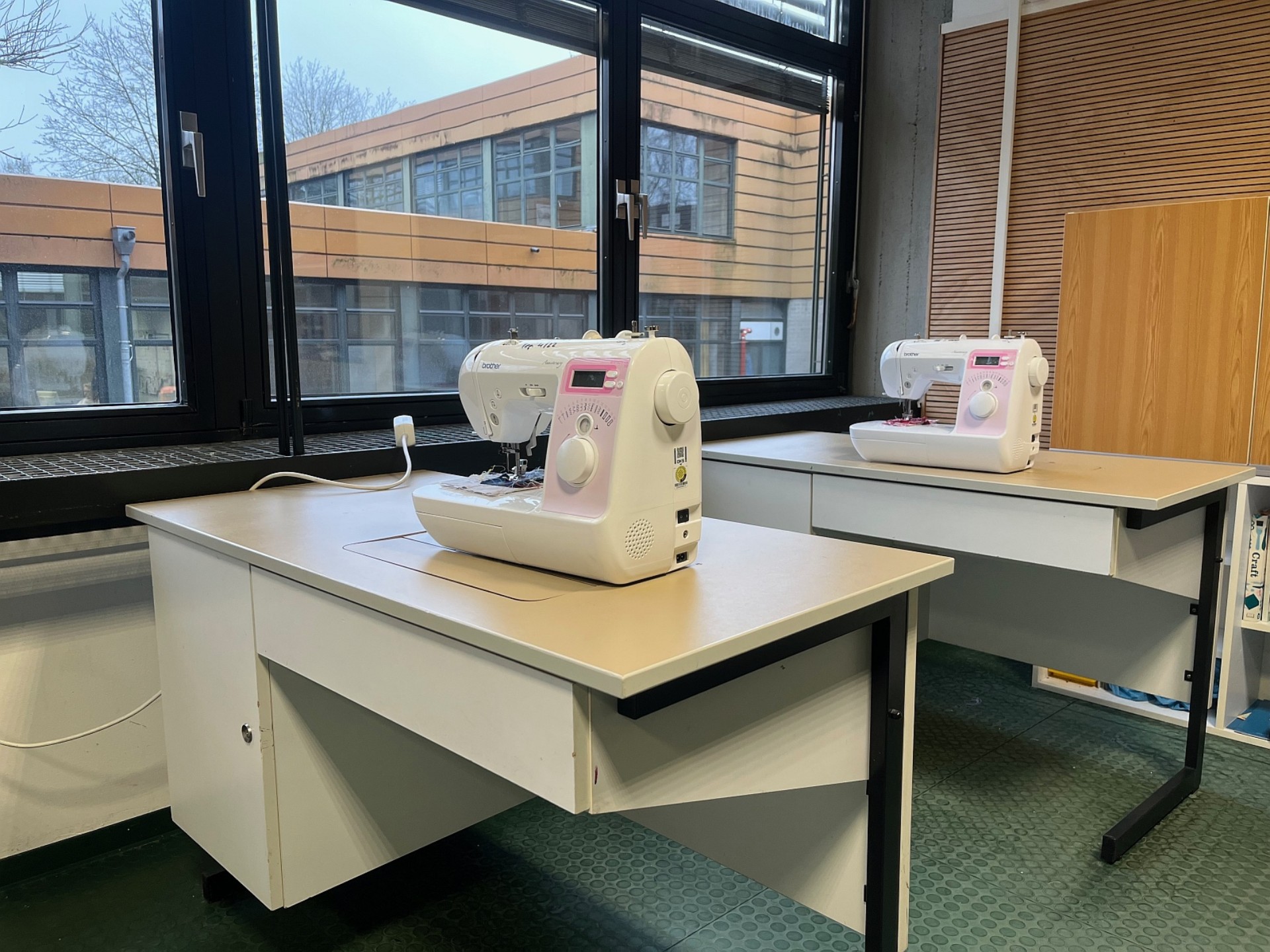 Ein heller Arbeitsraum mit zwei weißen Arbeitstischen, auf denen jeweils eine moderne Nähmaschine steht. Der Blick durch die Fenster zeigt ein gegenüberliegendes Schulgebäude.