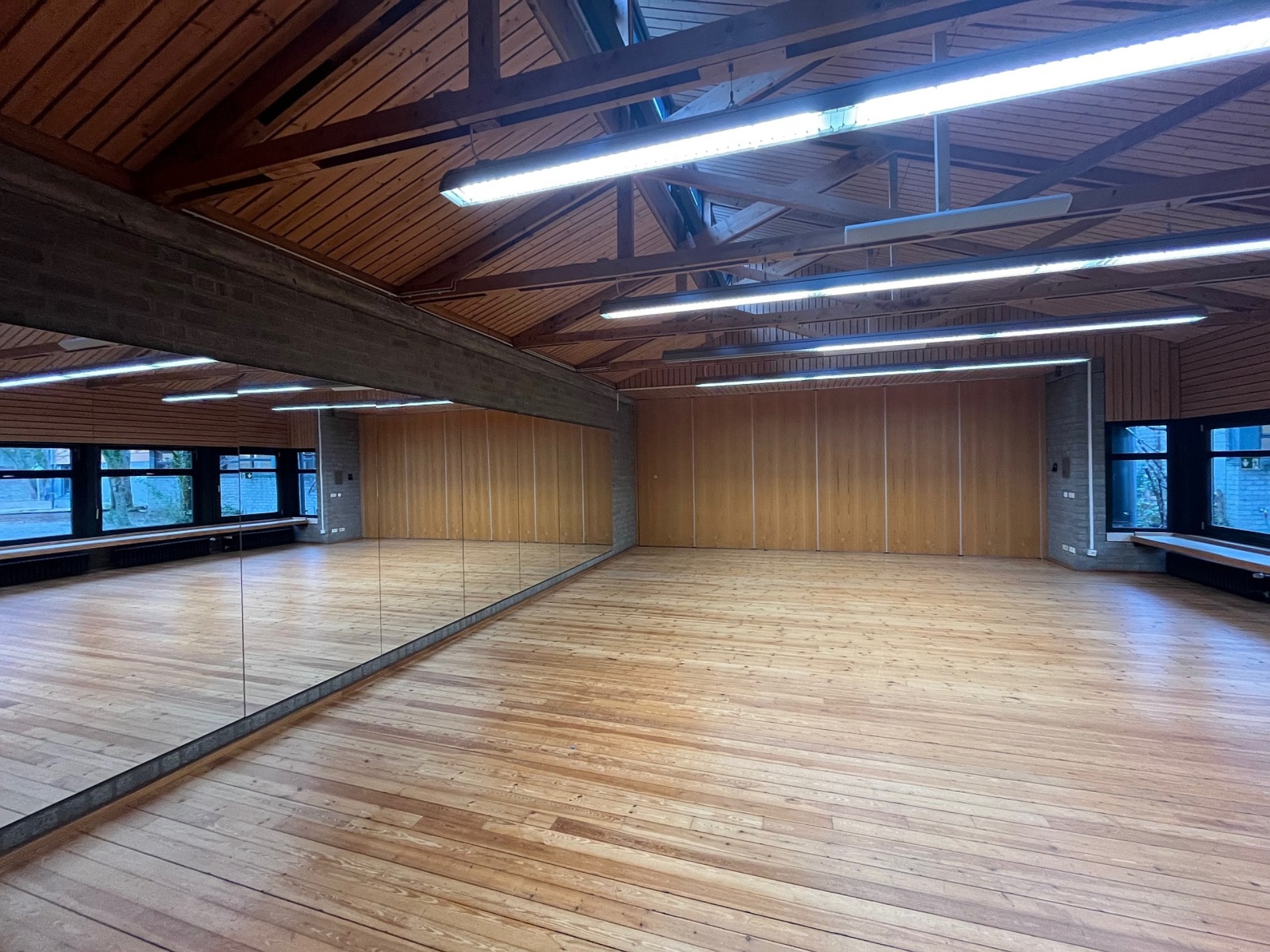 Ein großer Mehrzweckraum mit hellem Schwingboden aus Holz und einer markanten Sichtbalkendecke. Eine komplette Längswand ist verspiegelt, was den Raum ideal für Tanz oder Theater macht.