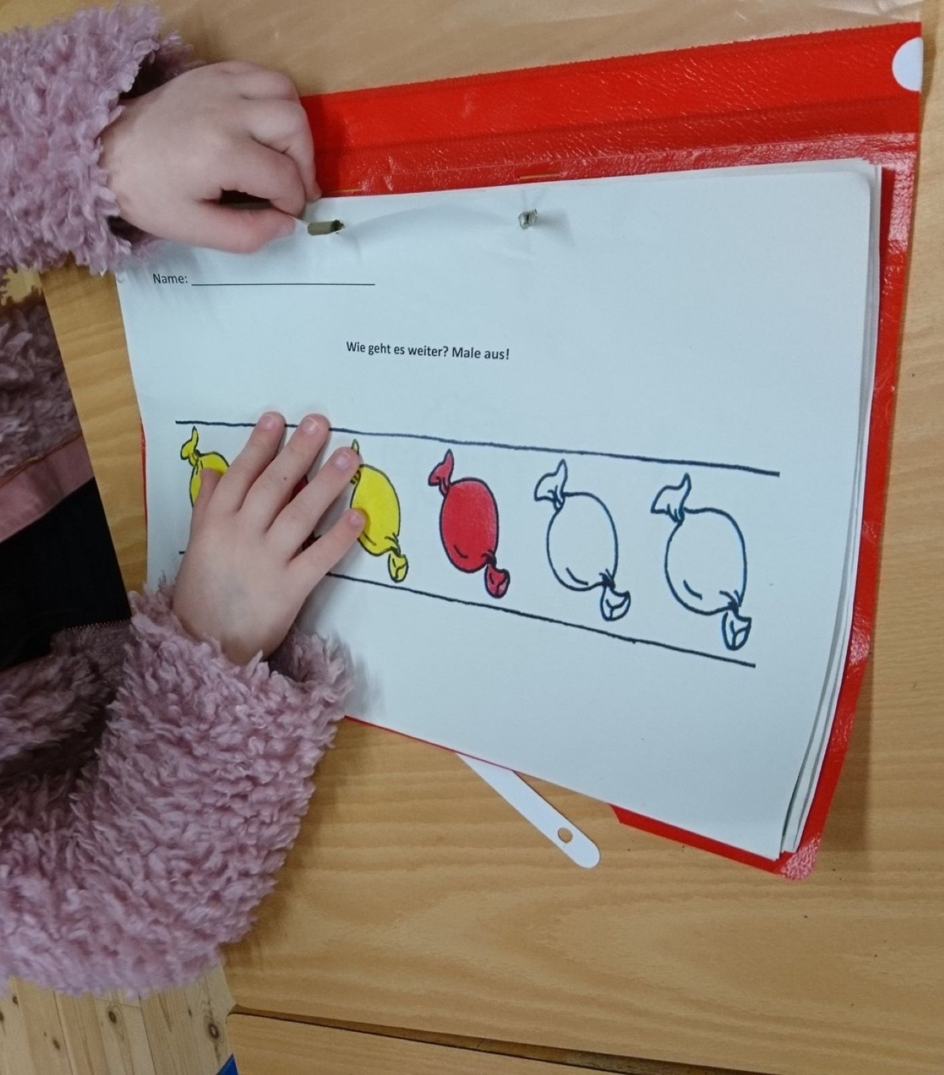 Auf dem Bild sieht man die Hände von einem Kind das an einem Tisch sitzt. Beide Hände liegen auf einem roten Schnellhefter der geöffnet auf dem Tisch liegt. Auf dem Arbeitsblatt im roten Schnellhefter ist ein Arbeitsblatt mit dem Arbeitsauftrag "Wie geht es weiter? Male aus!" zu sehen. Dadrunter ist eine Reihe 6 Bonbons. 4 davon sind abwechselnd gelb und rot angemalt. Zwei Bonbons müssen noch angemalt werden.