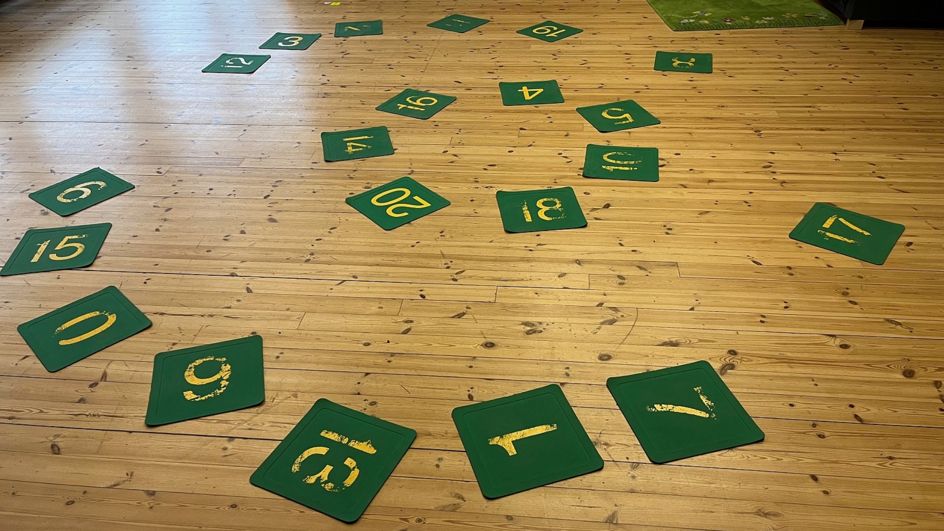 Ein heller Gruppenraum mit Holzboden, auf dem grüne Matten mit gelben Zahlen von 0 bis 20 in einer großen Spirale ausgelegt sind, um spielerisch den Zahlenraum zu erkunden