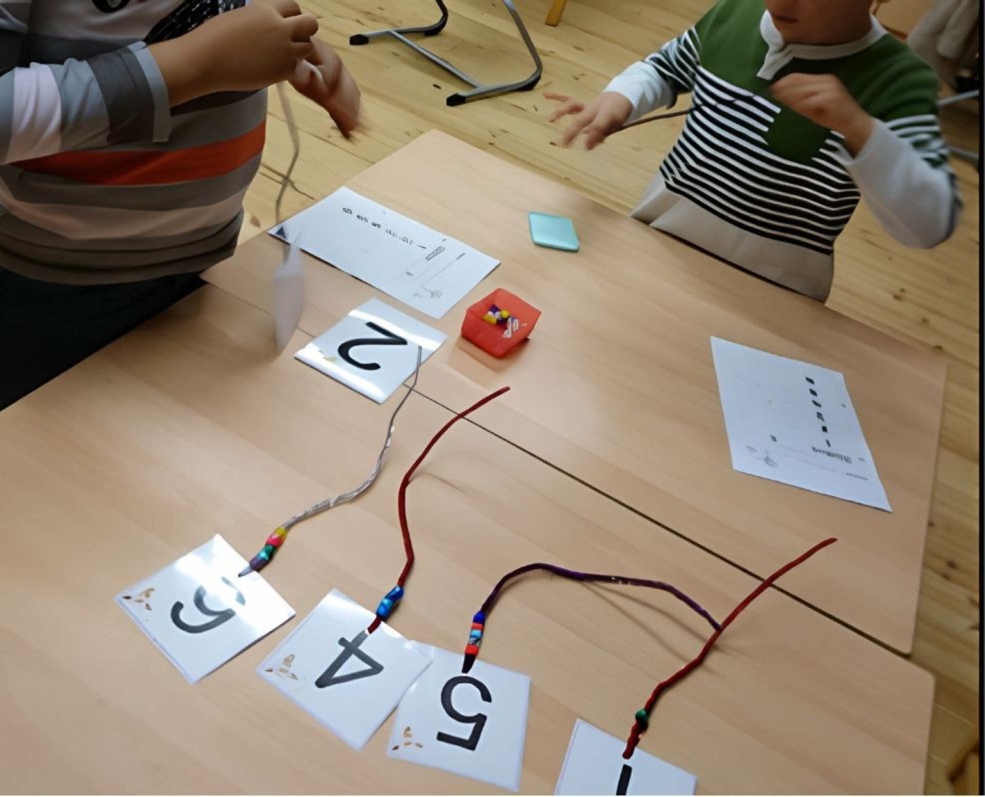 Auf dem Bild sieht man einen Tisch an dem zwei Kinder stehen. Auf dem Tisch liegen verschiedene kleine weiße Blätter mit Zahlen (1-6) drauf. An den Zahlen sind Pfeifenputzer mit Perlen. Die Kinder fädeln die Perlen auf.